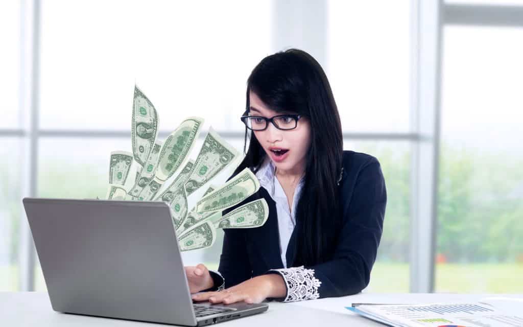 An excited woman looking at her laptop with money coming out of it