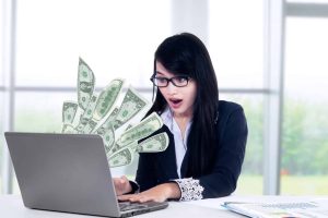 An excited woman looking at her laptop with money coming out of it